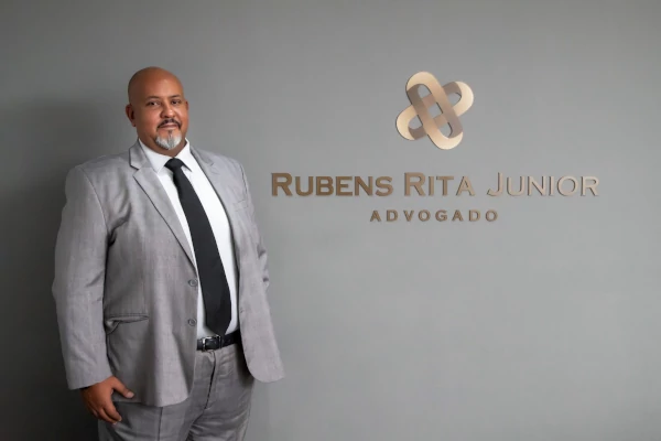 Dr. Rubens Rita Junior de pé ao lado da logomarca do escritório em uma parede cinza